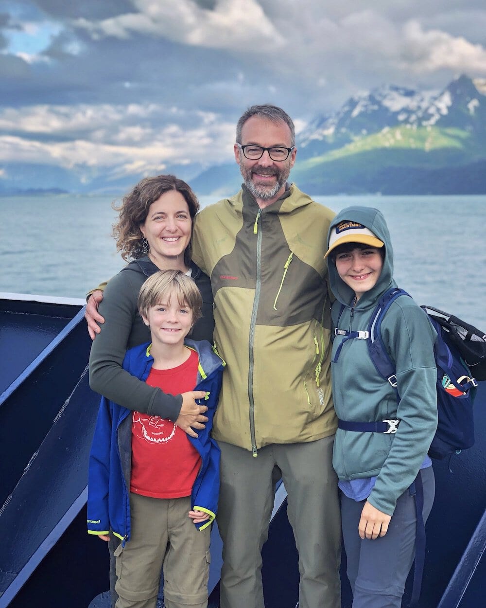 Our family of four smiling on the Aurora ferry from Whittier to Valdez with a scenic backdrop of mountains and overcast skies, portraying a moment of joy during our adventure.
