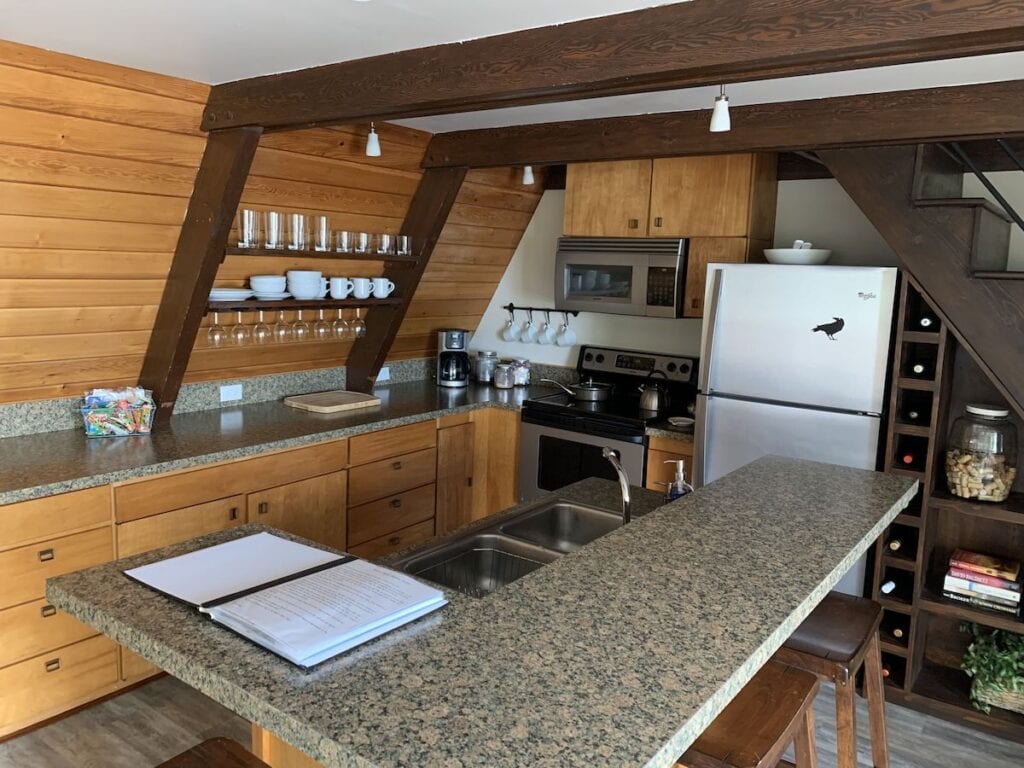 Modern kitchen inside one of the cozy hotels in Sitka Alaska, featuring wood-paneled walls, granite countertops, open shelving with glassware, and stainless steel appliances. A wine rack and rustic beams add a warm, cabin-style charm perfect for extended stays.