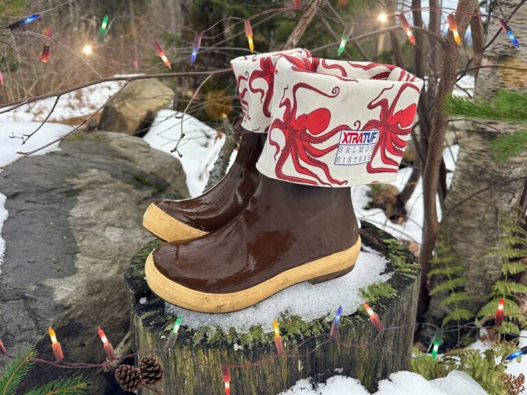 A pair of brown XTRATUF boots with light tan soles and cream-colored cuffs featuring a red octopus print, branded with a Salmon Sisters patch. The boots sit on a snow-dusted tree stump surrounded by moss, with holiday string lights draped across bare branches and pinecones nestled nearby, evoking a festive outdoor winter scene in Alaska. These are at the top of our Alaska Holiday Gift Guide!